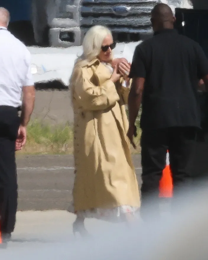 Lady Gaga holding her newborn daughter while wearing a tan trench coat and sunglasses, walking with two men.