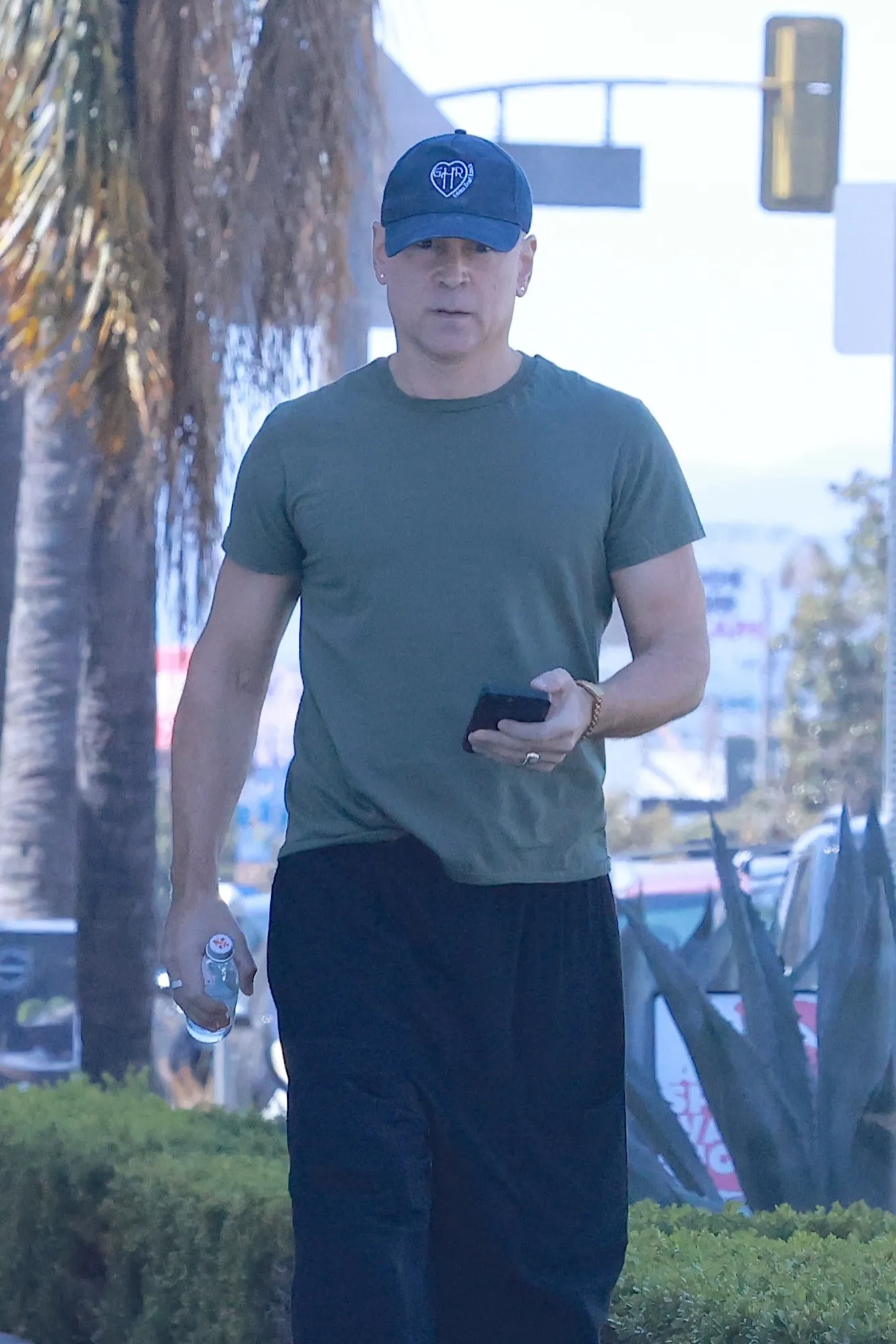 Colin Farrell with a shaved head and a blue baseball cap walks outside.