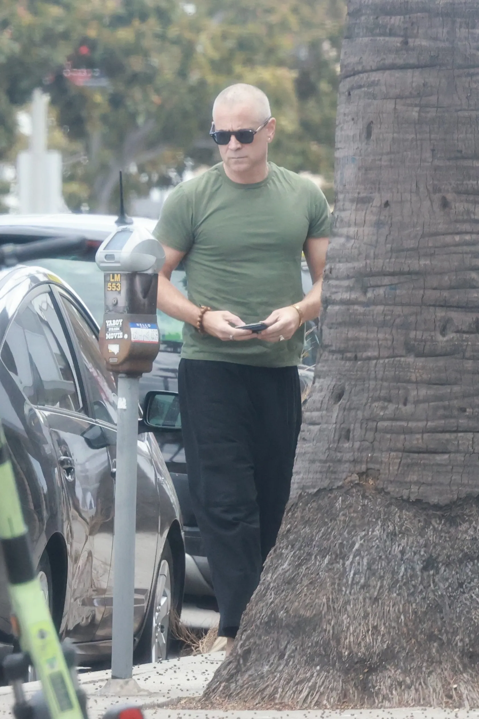 Colin Farrell with a newly shaved head, wearing a green t-shirt and black pants, walking outdoors.