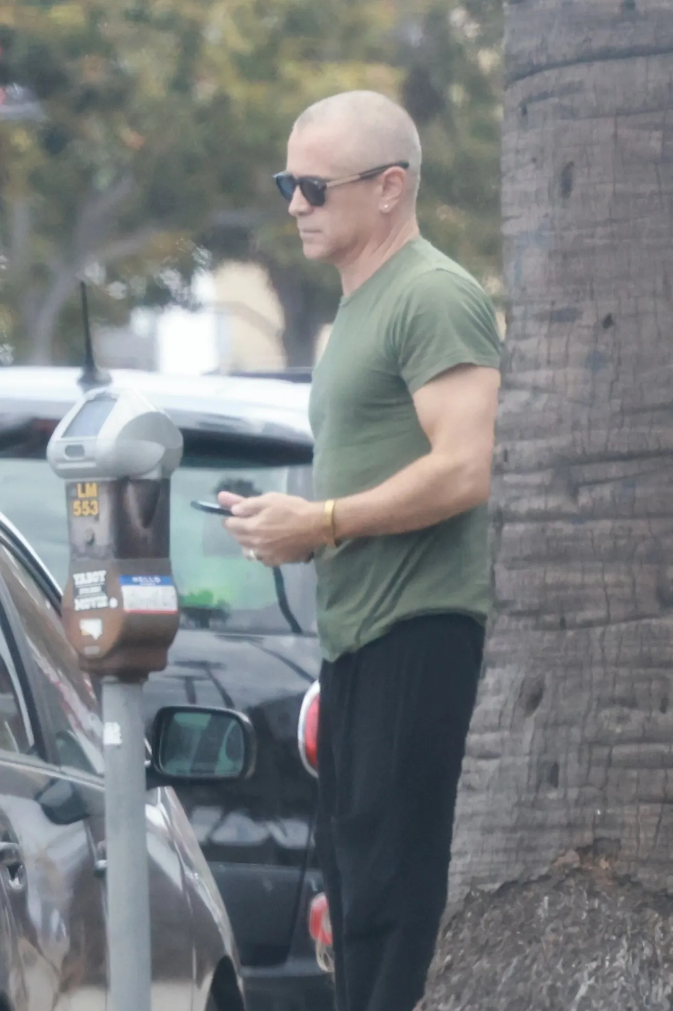 Colin Farrell with a shaved head and sunglasses, wearing a green t-shirt and dark pants, holding a phone.