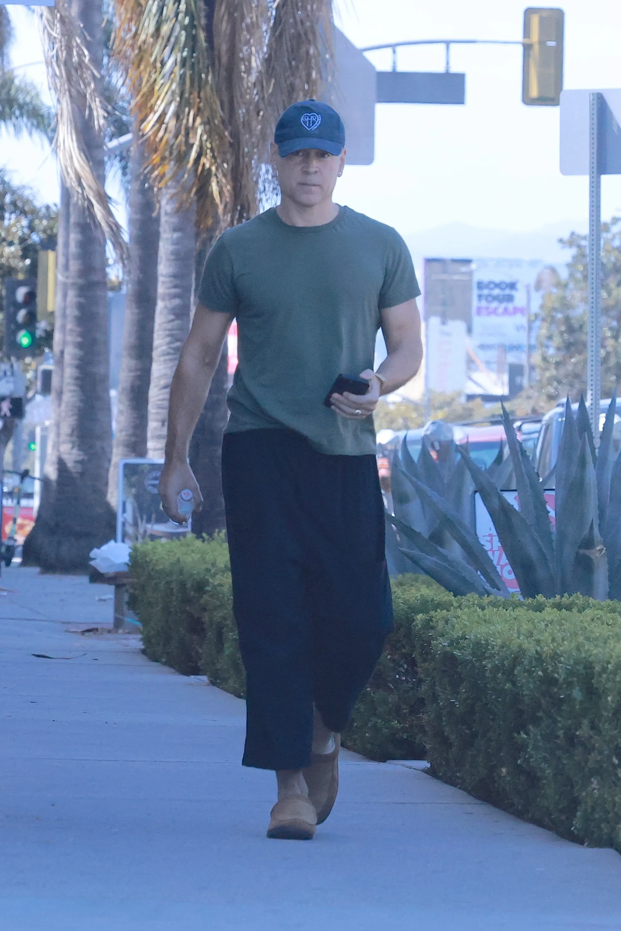 Colin Farrell walking outdoors with a shaved head, wearing a green t-shirt, dark pants, and a blue cap.