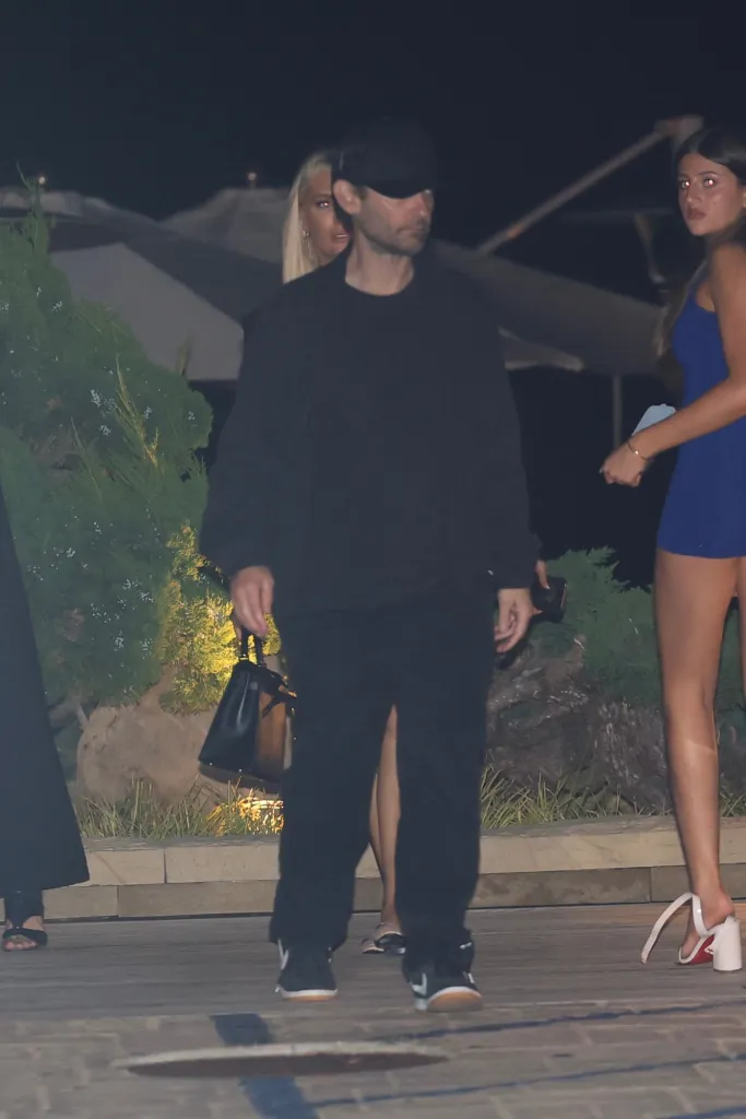 Tobey Maguire in a black hat and clothing walking with a woman in a blue dress and a blonde woman.