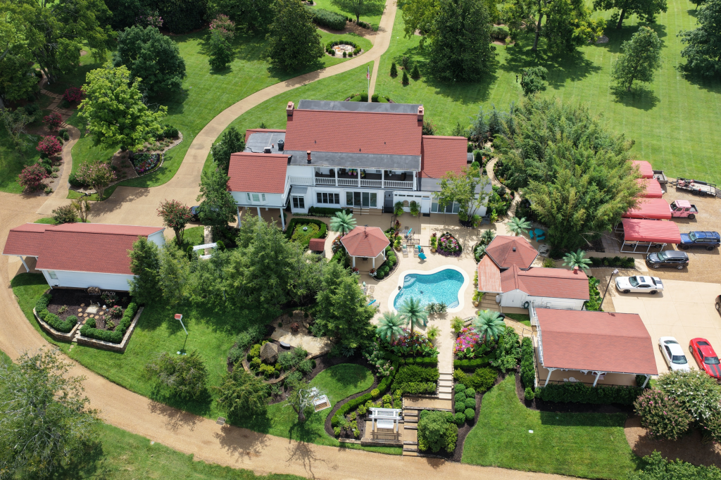 Aerial view of Dolly Parton's private Tennessee compound, featuring a white house with a red roof, a swimming pool, and multiple outbuildings surrounded by lush greenery and driveways.