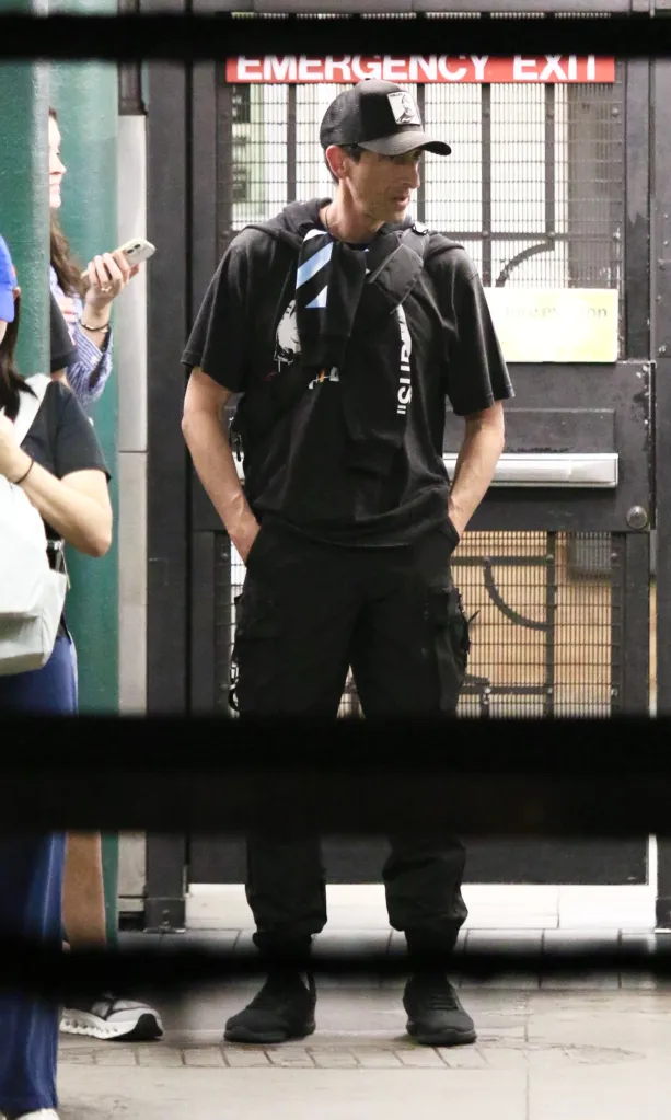 Adrien Brody in a black outfit and baseball cap stands on a New York City subway platform with a friend.