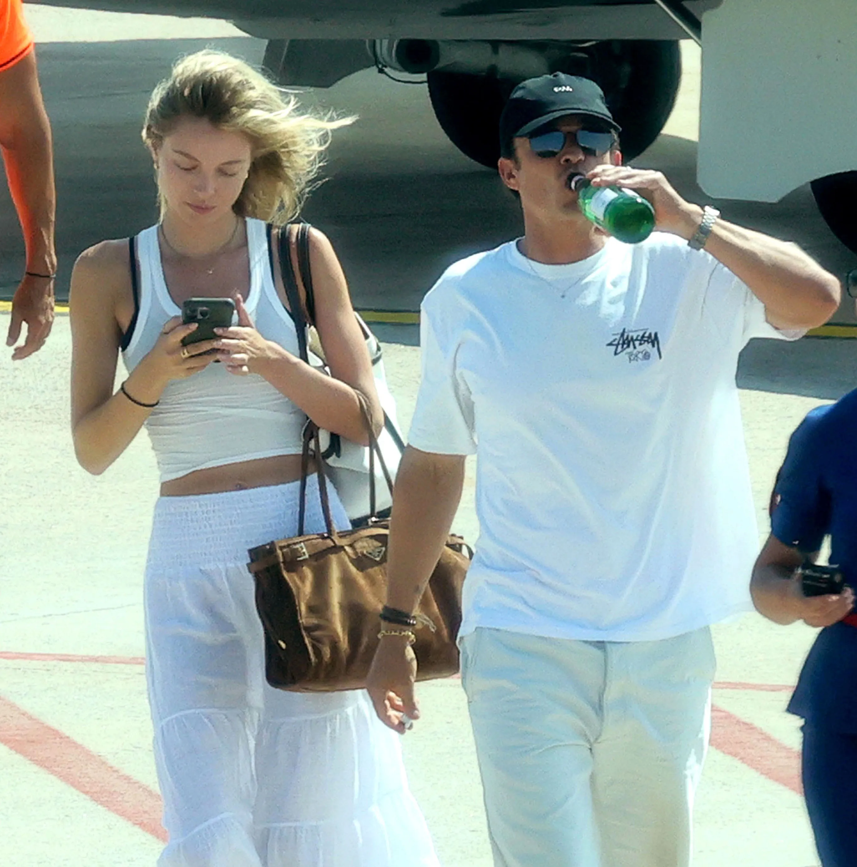 Orlando Bloom in a white t-shirt and shorts drinks from a green bottle next to Luisa Laemmel, who is wearing a white tank top and skirt and looking at her phone.