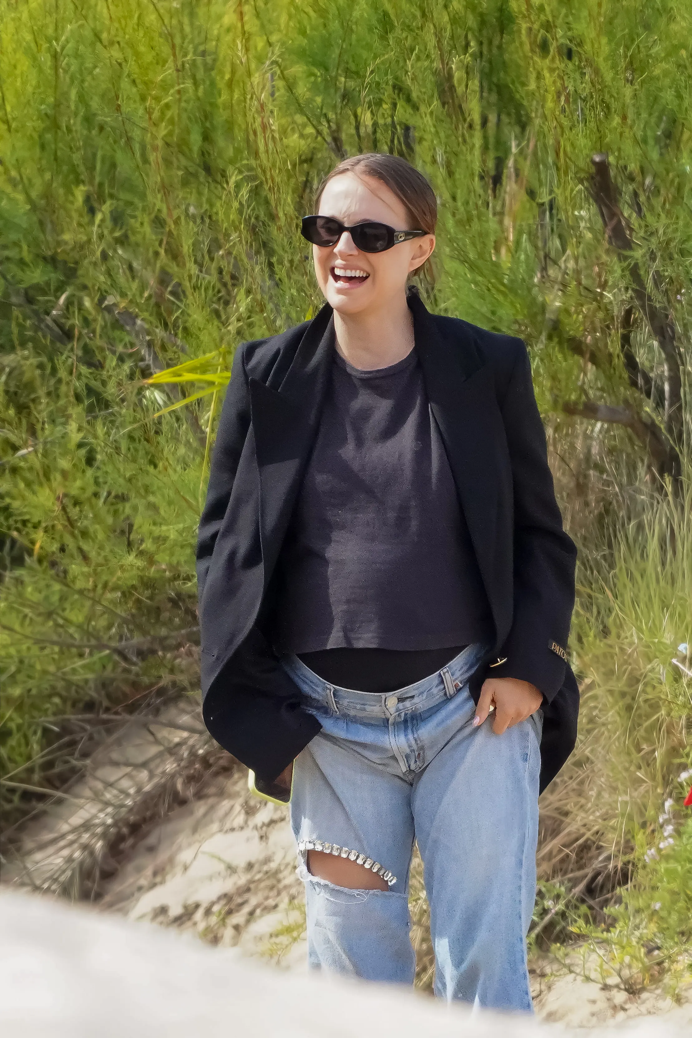 Natalie Portman laughing outdoors in a black blazer, light wash ripped jeans, and sunglasses.
