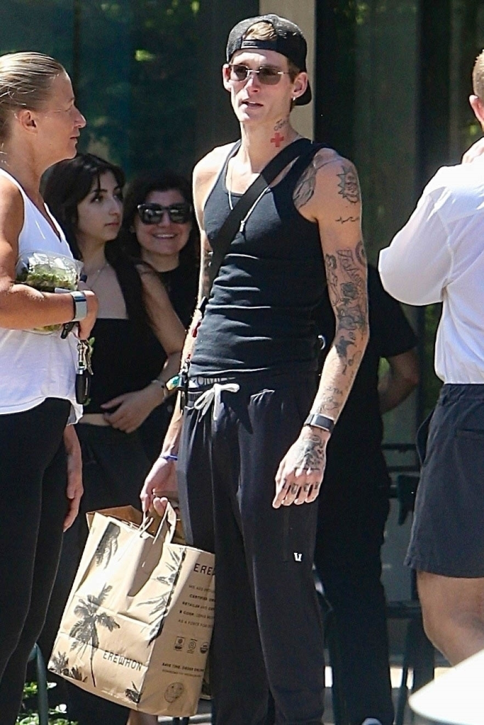 Presley Gerber, wearing a black tank top and matching sweatpants, holding a paper shopping bag outside of Erewhon, surrounded by other people.