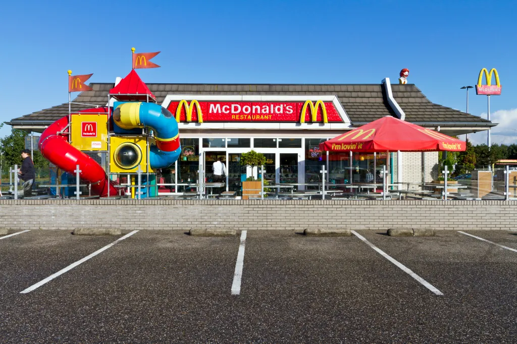 An exterior view of a McDonald's restaurant with a red and yellow playground, red umbrellas on a patio, and a yellow McDonald's sign.