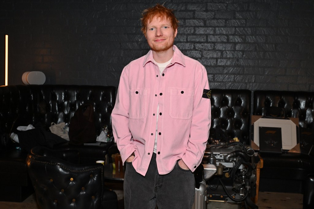 Ed Sheeran in a pink shirt and black pants, smiling at the camera.
