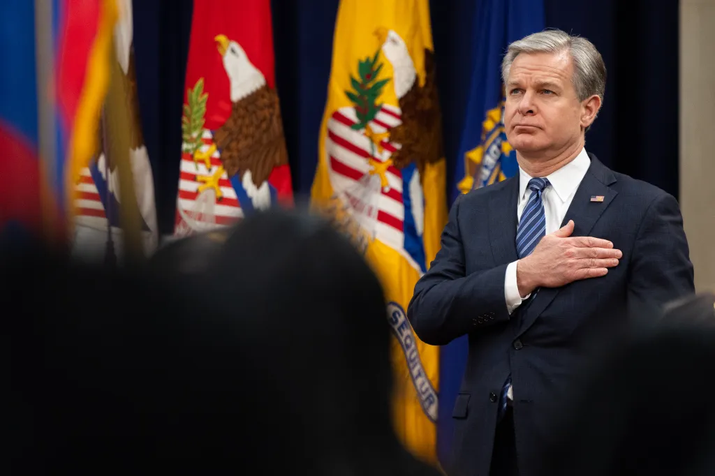 FBI Director Christopher Wray stands with his hand over his heart at Attorney General Merrick Garland's farewell ceremony.