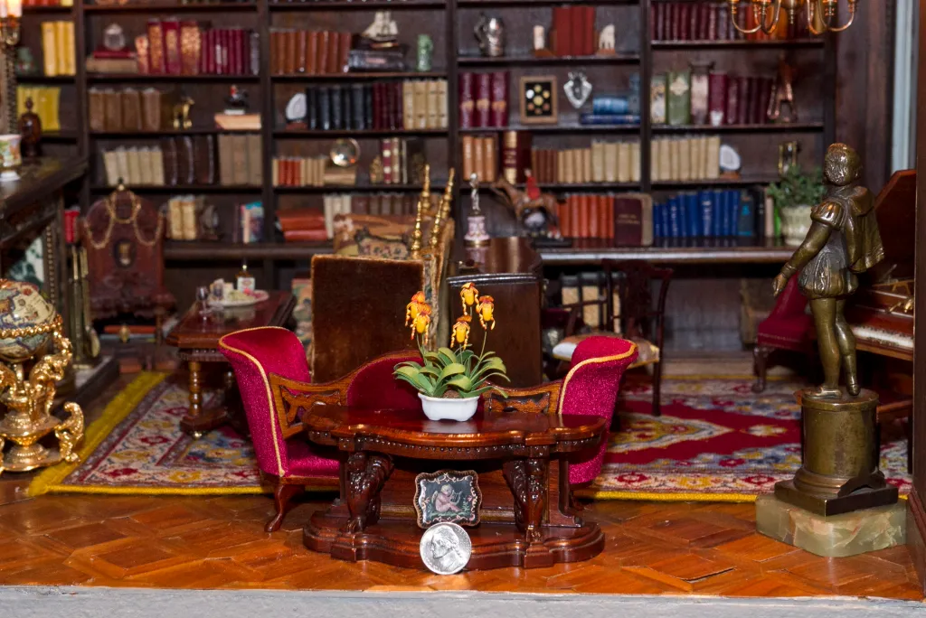 The inside of the Astolat Dollhouse Castle, with a small table with a flower, red chairs, and a bookshelf in the background.