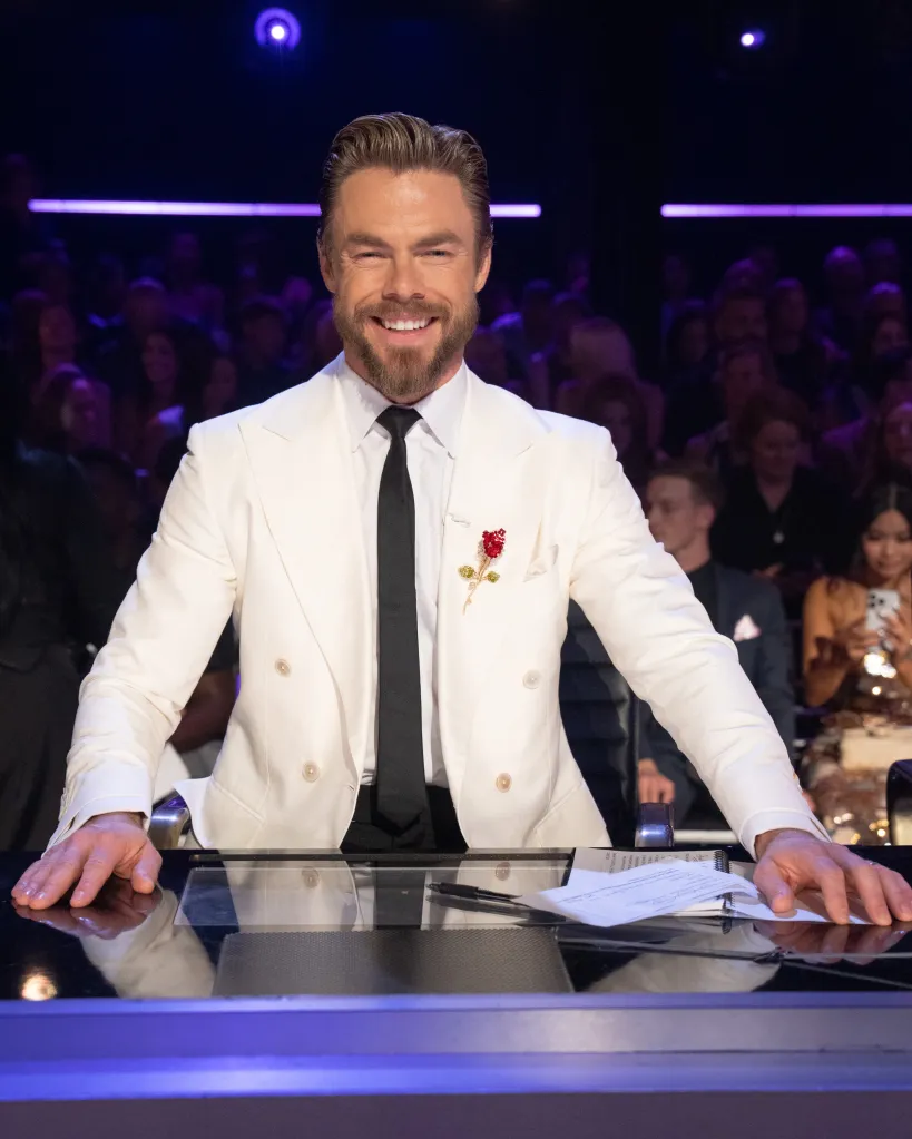 Derek Hough smiling at the camera from the judge's table.