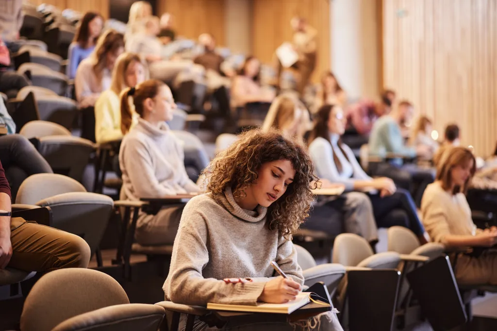 A female college student writes an exam in a lecture hall with her classmates in the background.
