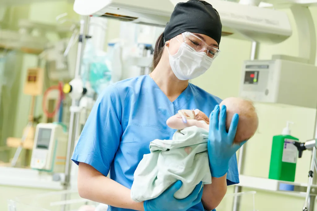 A female nurse wearing a mask and goggles holds a newborn baby at a neonatal resuscitation center.