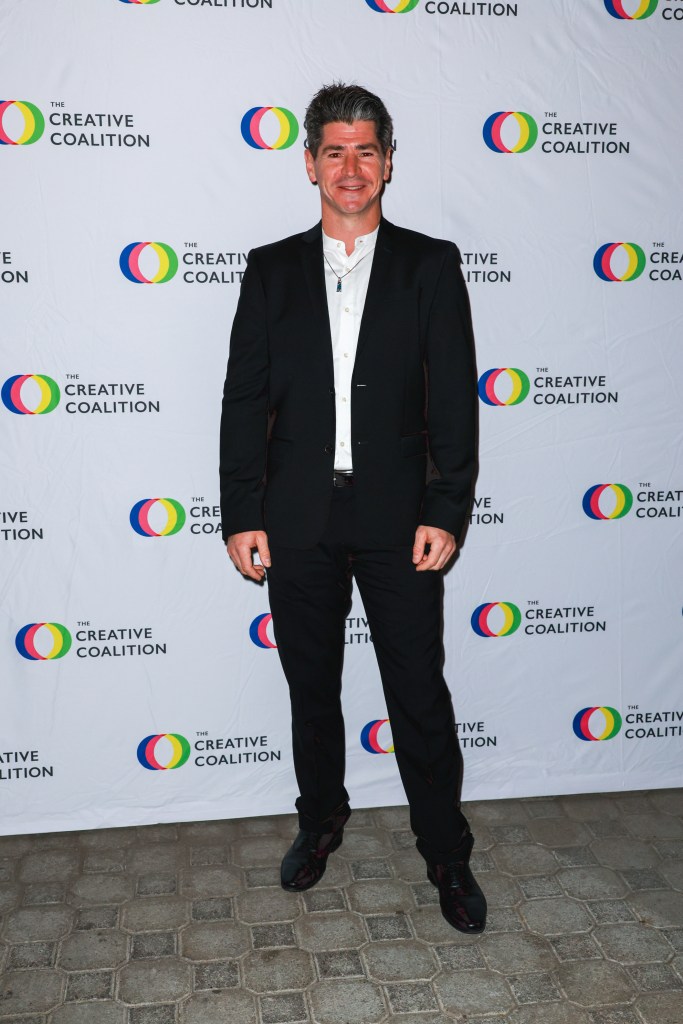 Michael Fishman posing in a black suit with a white shirt and necklace in front of a white backdrop with 