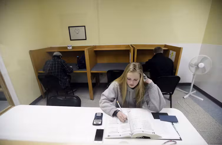 A white female student with long blonde hair sits at a desk and looks over an open book, with a calculator next to her.