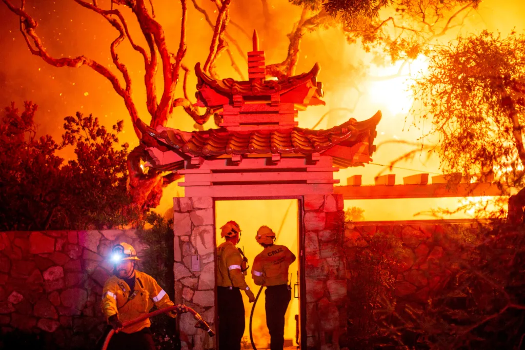 Firefighters battling the Palisades Fire, with a firefighter in the foreground holding a hose and two others visible through a gateway.