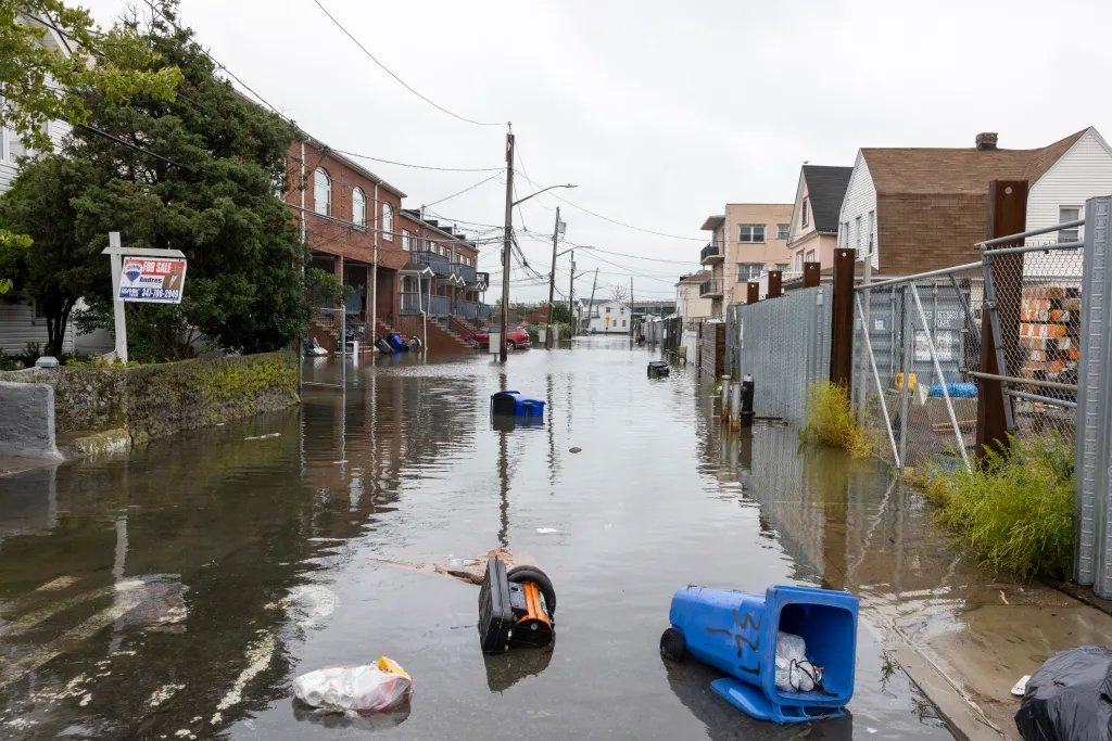 Flooded street in Far Rockaway, Queens, with trash cans and debris floating in the water.
