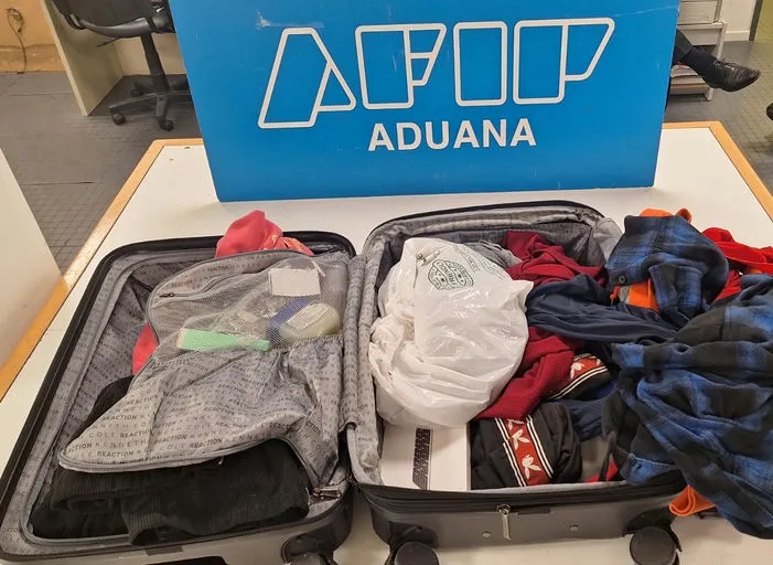 An open suitcase on a table with clothes and a white plastic bag, next to a blue sign with 