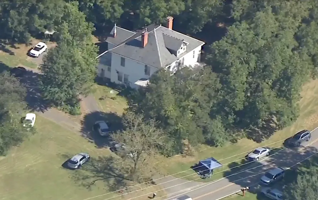 Aerial view of a white house surrounded by trees, with several cars parked on a nearby road and a blue tent set up on the shoulder.