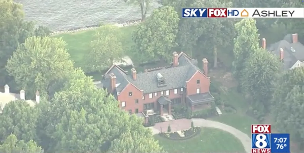 Aerial view of a large, red brick house with a dark roof surrounded by dense green trees and a body of water in the background.