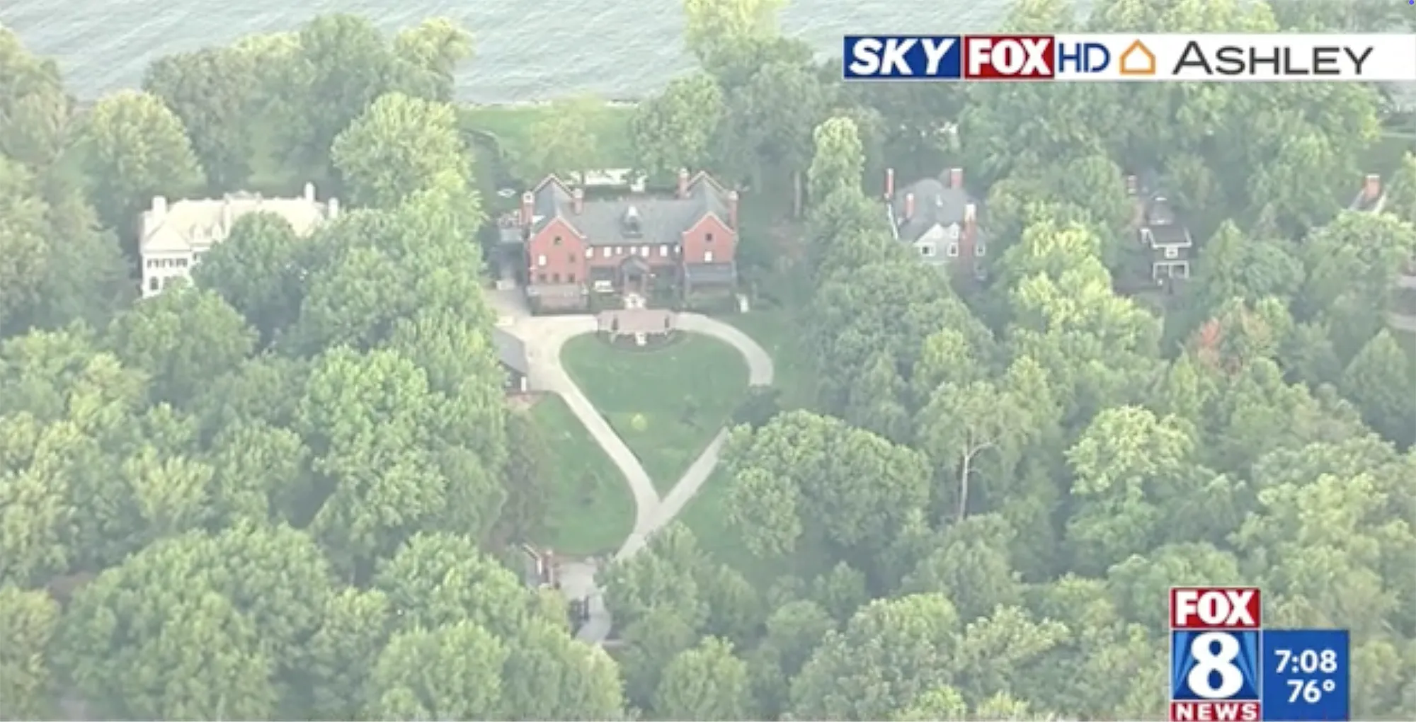 An aerial view of a red brick mansion surrounded by green trees, with a heart-shaped driveway.