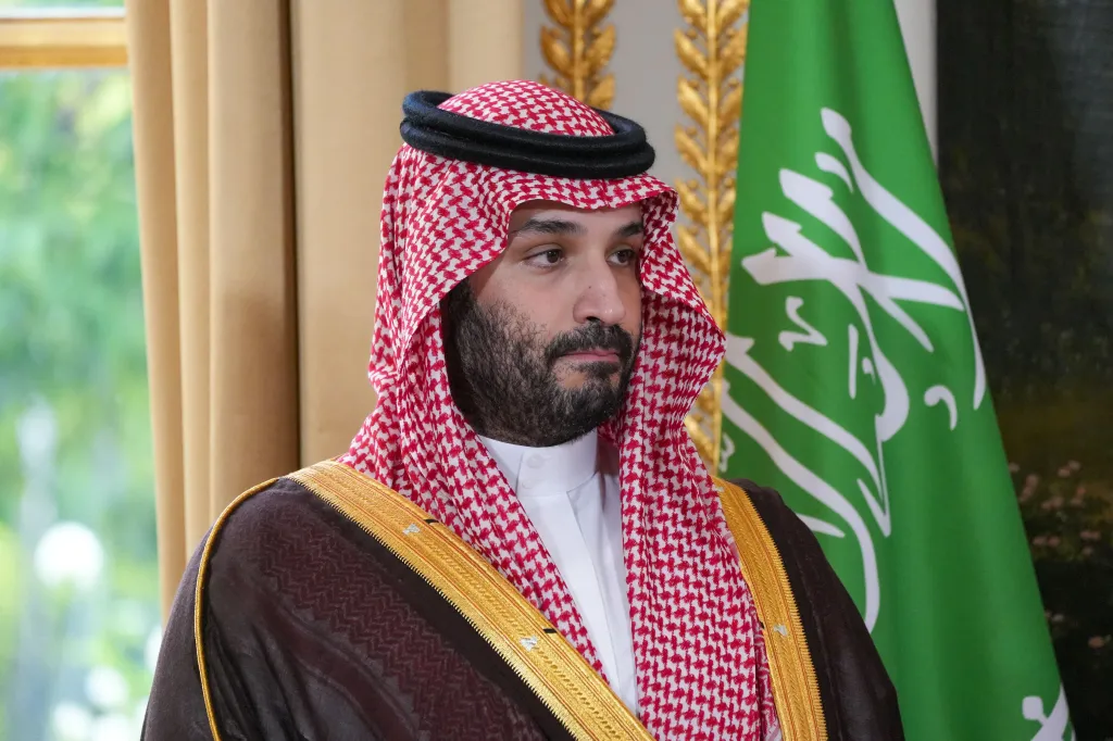 Saudi Crown Prince Mohammed bin Salman wears traditional Saudi attire, looking off to the right, with a Saudi flag visible in the background.