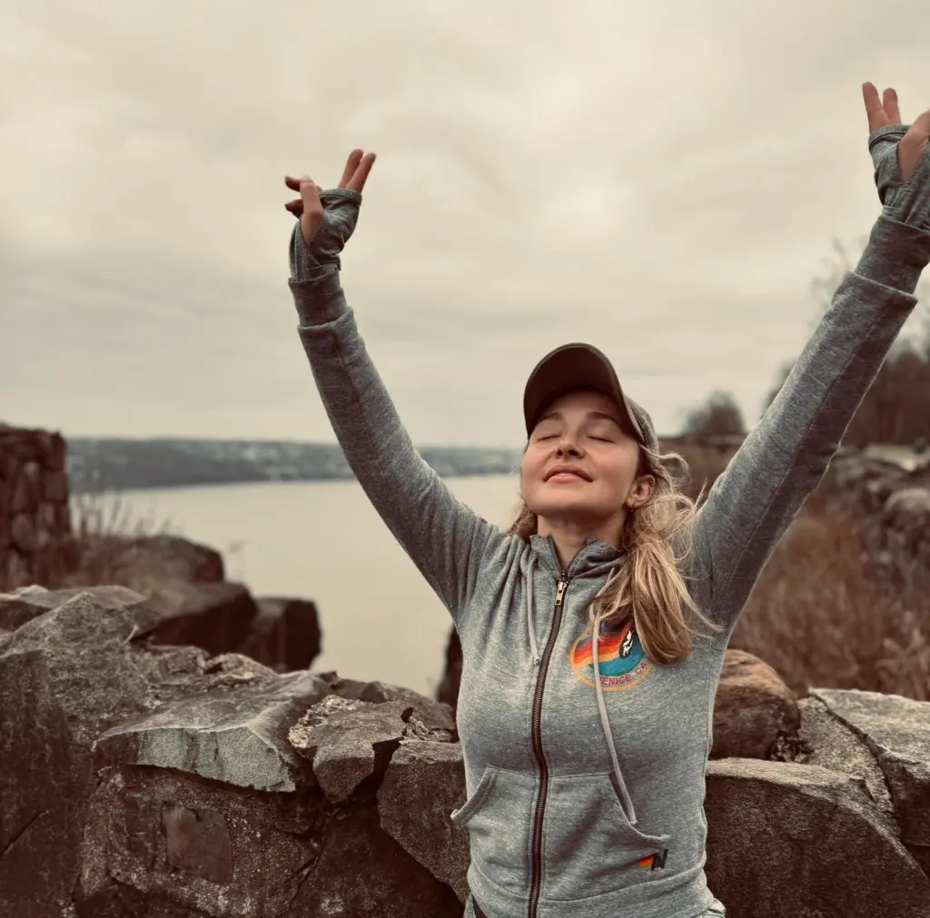 Hayden Panettiere with her arms raised in celebration on a rocky overlook with a body of water in the background.
