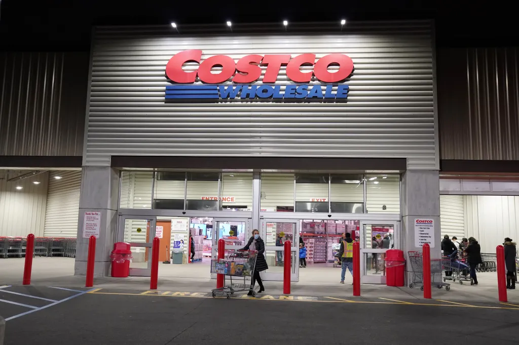 A nighttime exterior view of a Costco store with its red and blue logo above the entrance, with several people entering and exiting.