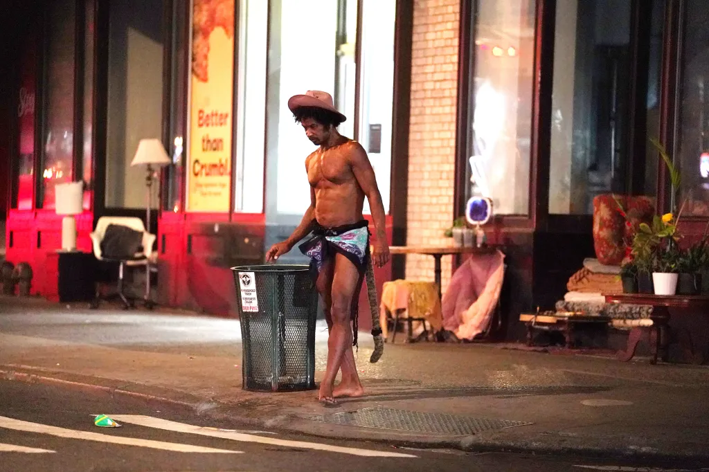A shirtless man Tiger in a hat and sarong stands next to a trash can on a city street at night, with furniture and lights in the background.