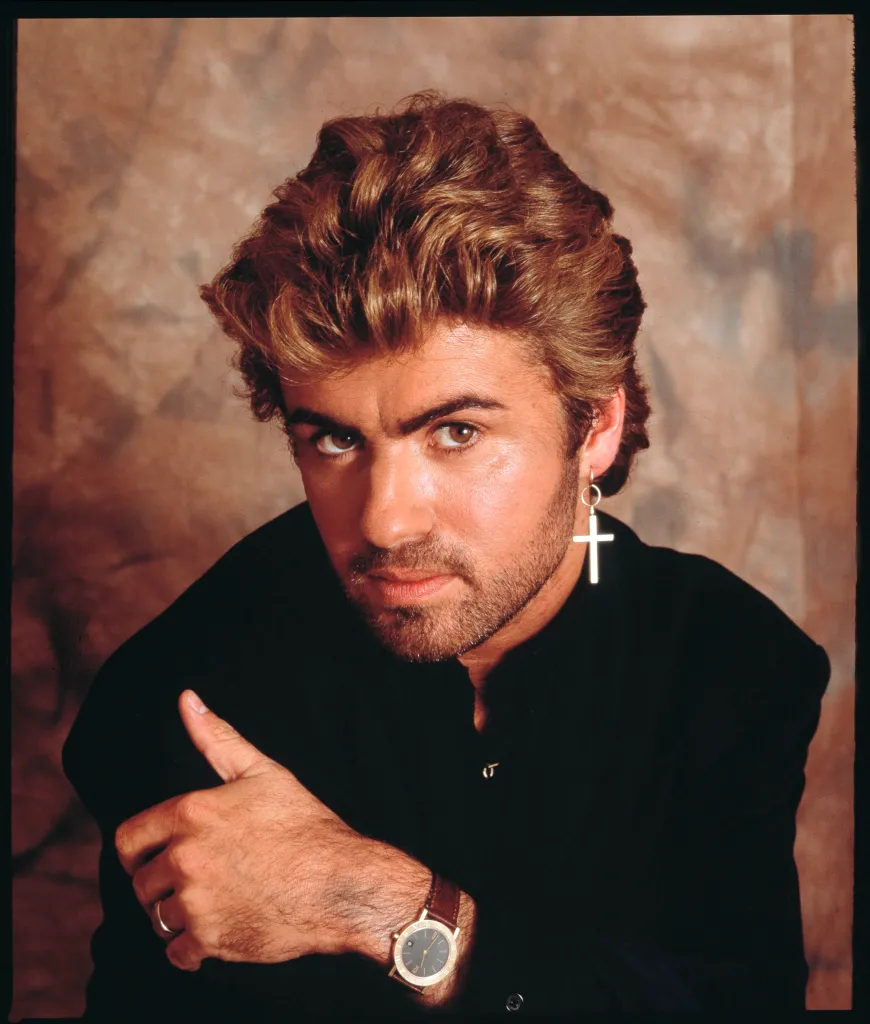 Studio portrait of George Michael in a black shirt with a cross earring.