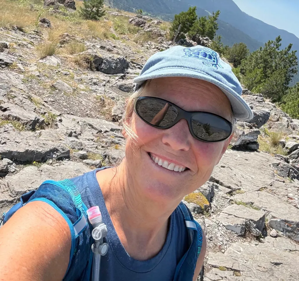 Mary Sorensen, smiling in sunglasses and a baseball cap, on a rocky mountain.