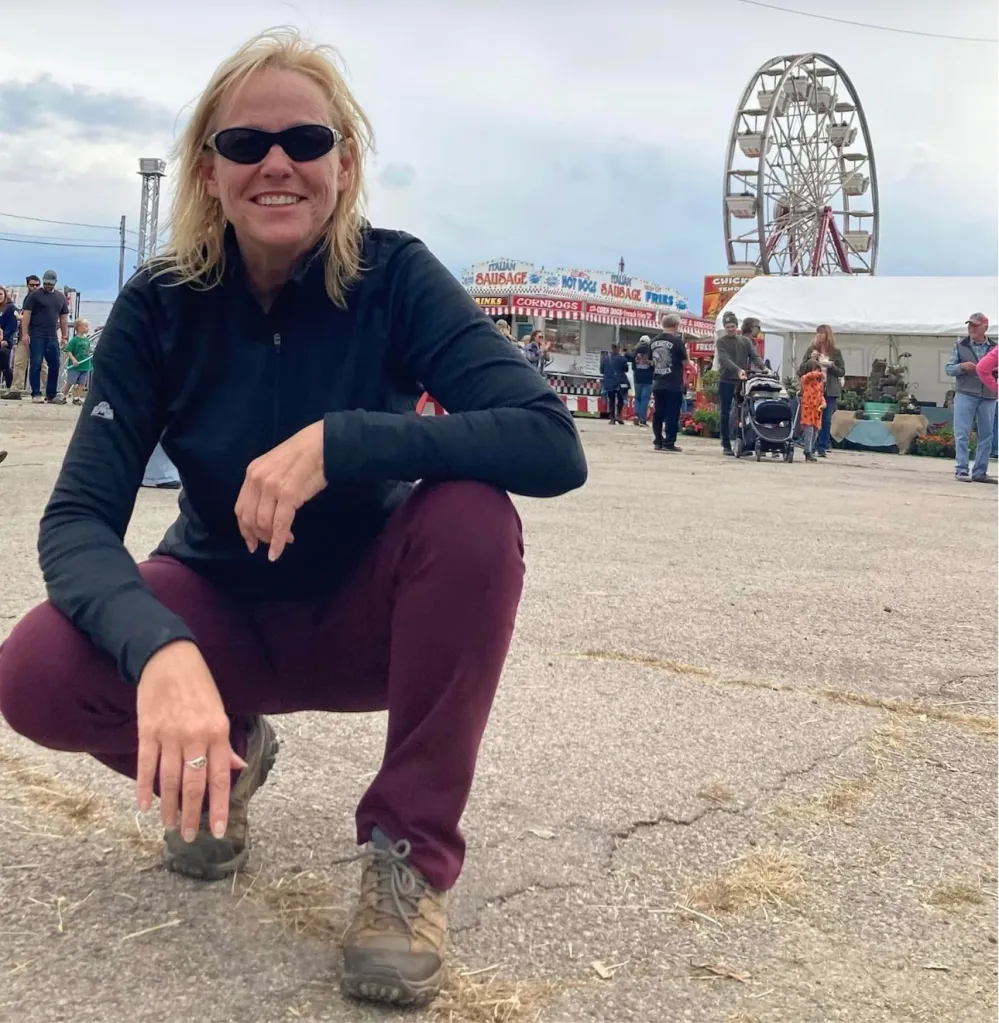 Mary Sorensen squatting and smiling at a fairground with a Ferris wheel and food stalls in the background.