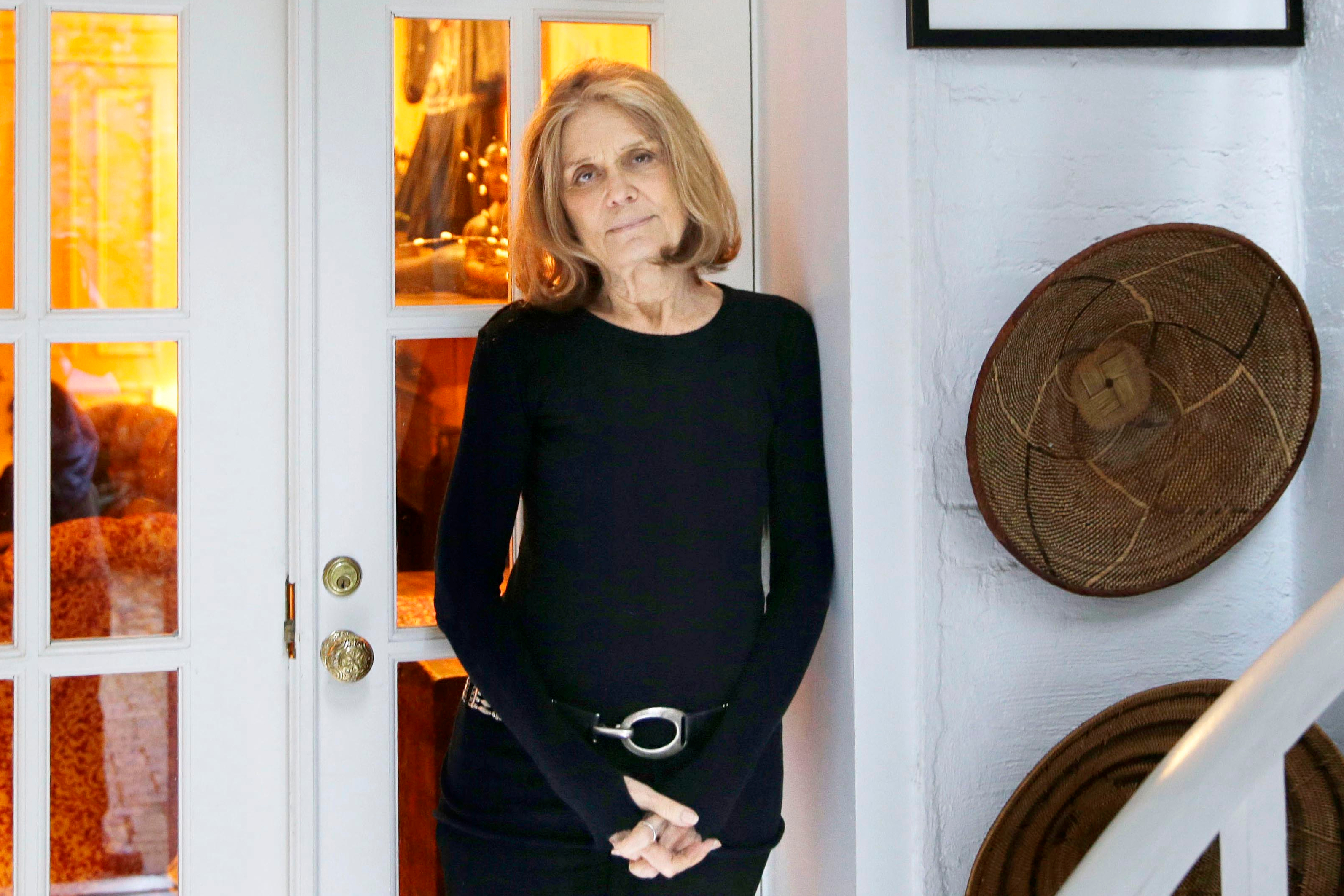 Gloria Steinem in a black dress, leaning against a white door with glass panels.