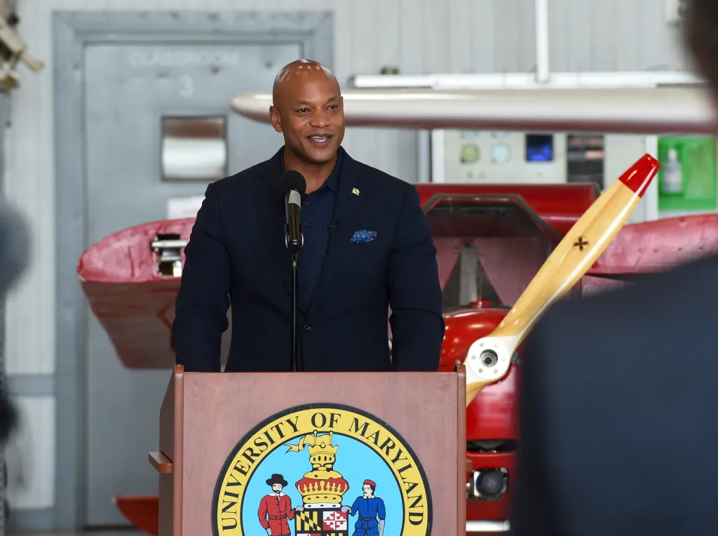 Governor Wes Moore speaking at a podium with a University of Maryland seal, in front of a red airplane with a propeller.