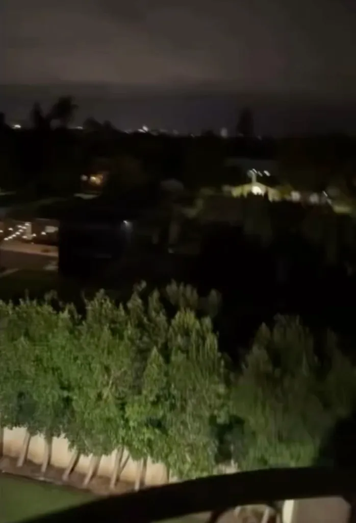 A nighttime view of a neighborhood from an elevated vantage point, showing lit houses, trees, and a dark sky.