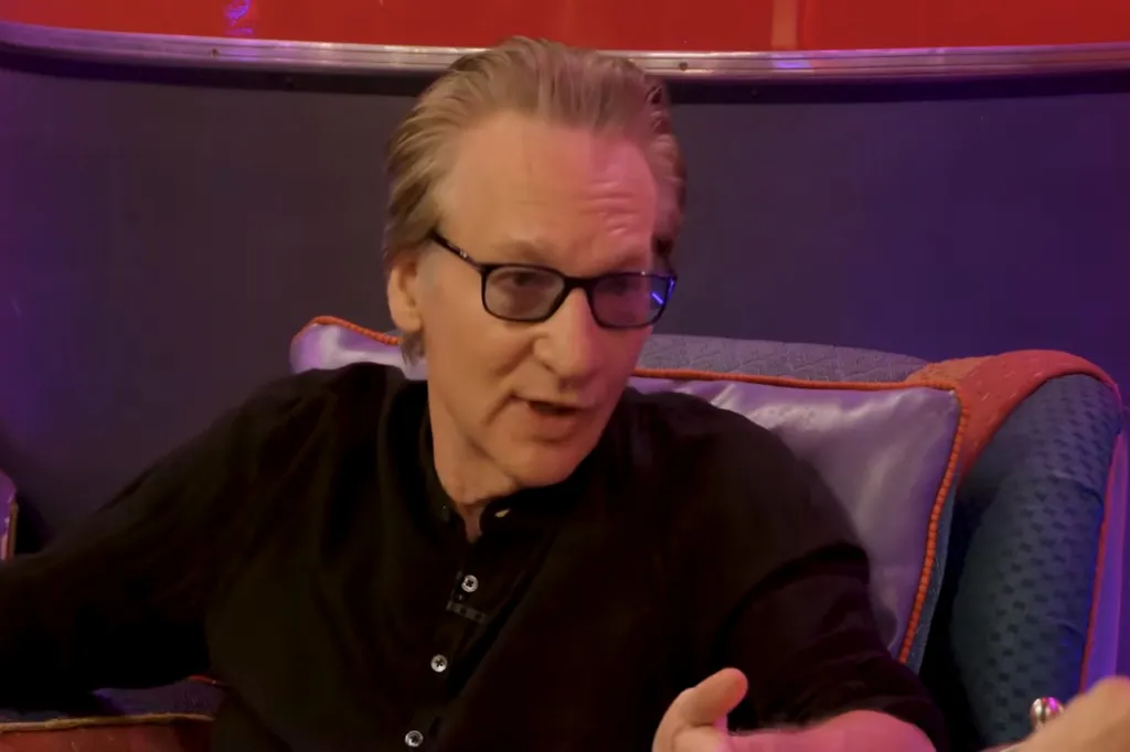 Bill Maher wearing black-rimmed glasses and a black shirt, speaking on his podcast
