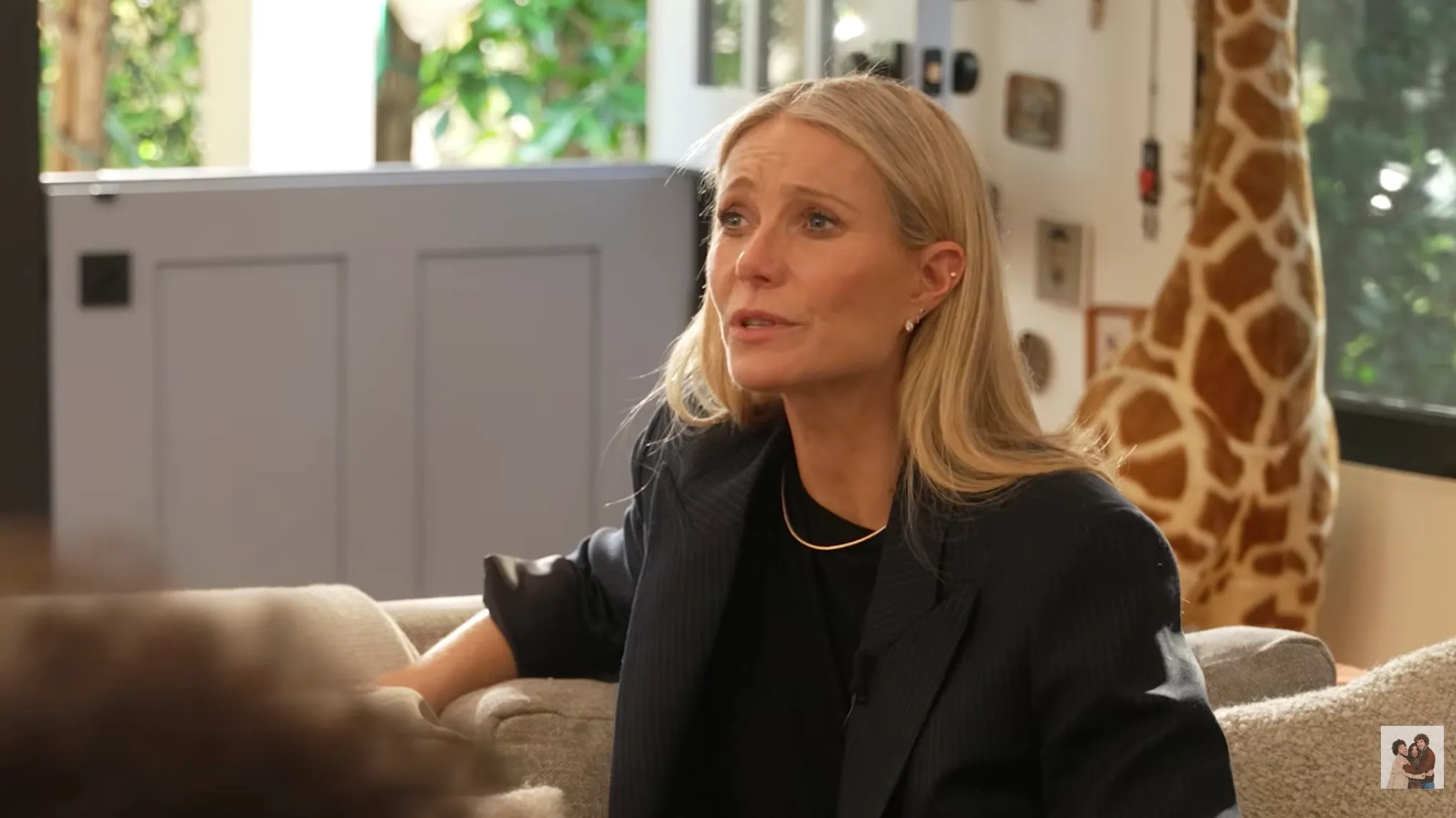Gwyneth Paltrow looking upset as she sits on a couch in a room with a giraffe statue in the background.