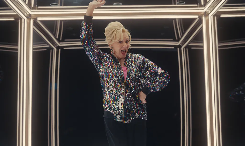 Jean Smart in a sequined jacket, posing with one arm raised in an illuminated box.