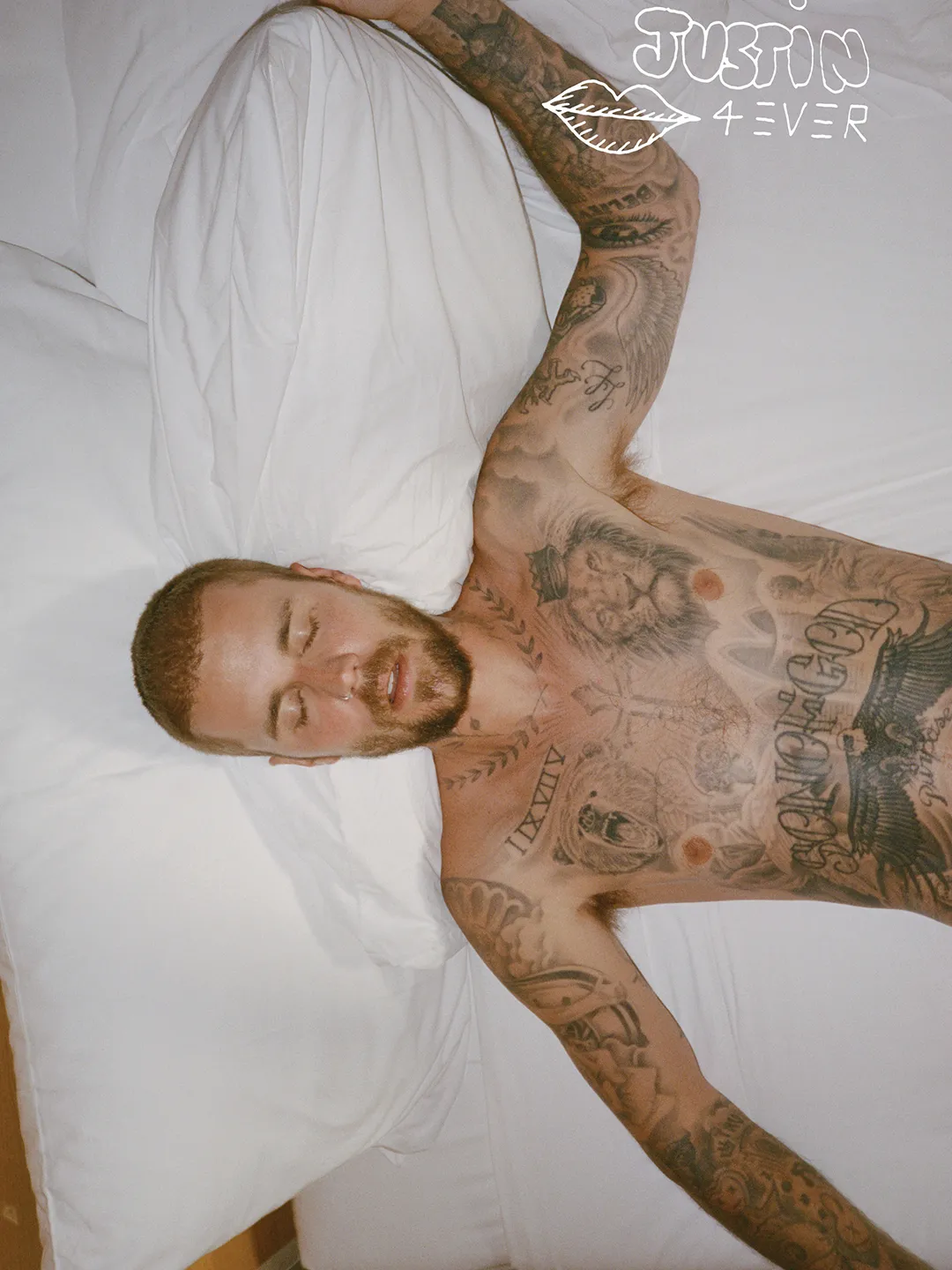 Justin Bieber lying shirtless on a white bed, covered in tattoos.