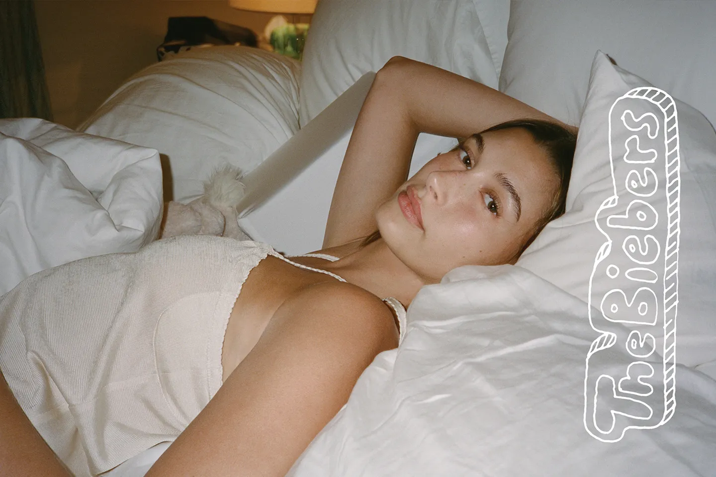 Hailey Bieber lying on white bedding with an arm under her head and 