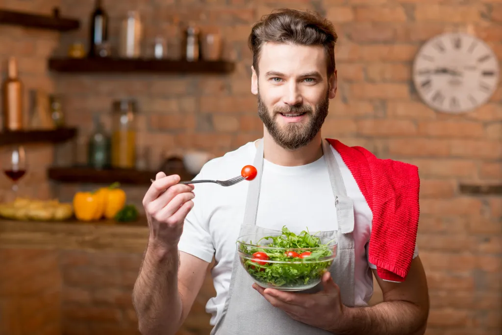 Smiling man in an apron holding a salad bowl and a fork with a cherry tomato.