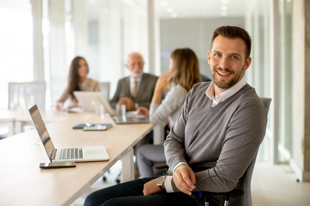A young businessman smiles at the camera with his teammates working in the background.