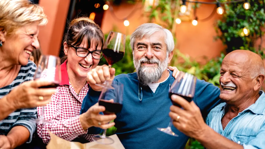 A group of four senior friends toasting with red wine, smiling and laughing at a dinner party.
