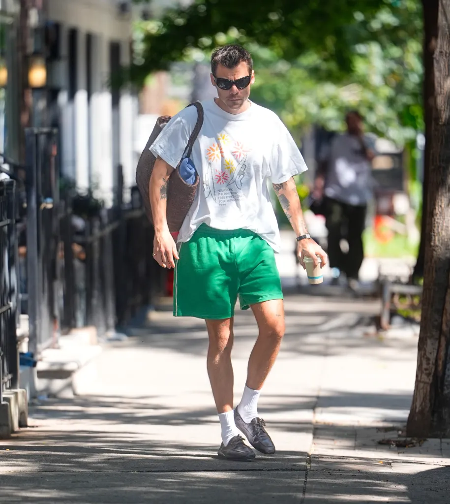 Harry Styles in sunglasses, a white graphic t-shirt, green shorts, and boat shoes, walking on a sidewalk while carrying a coffee and a bag over his shoulder.