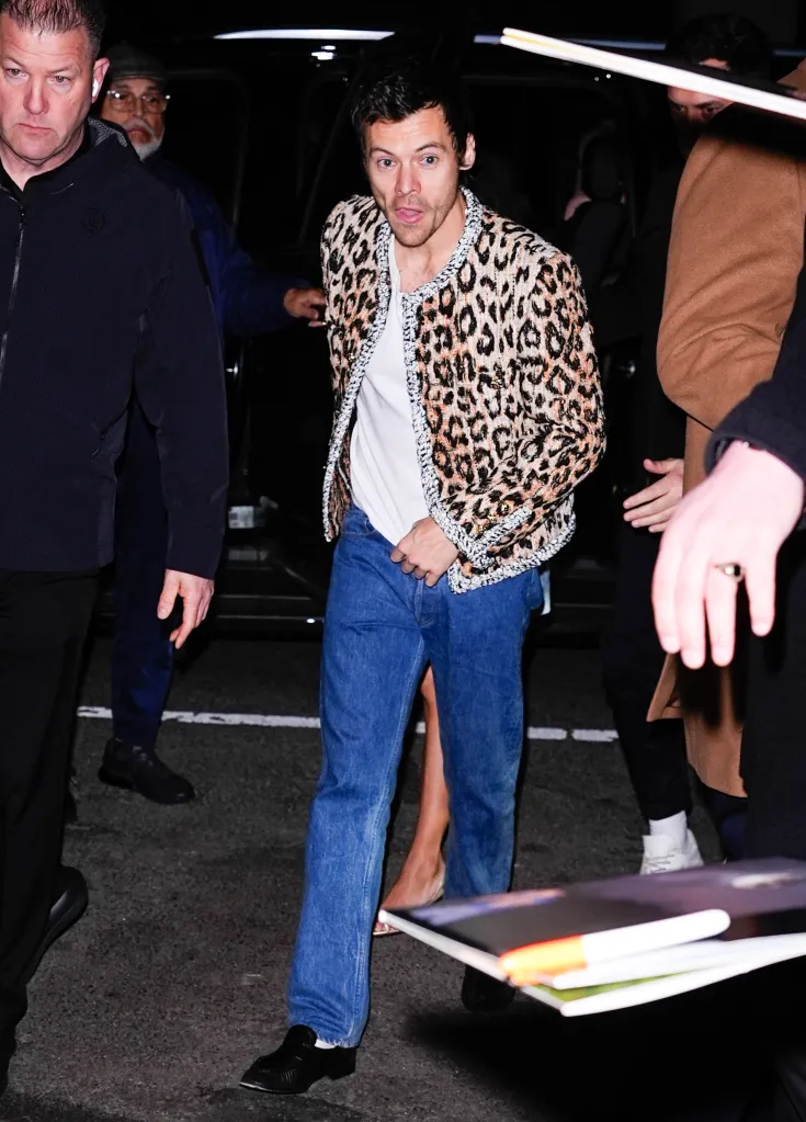 Harry Styles arriving in New York City, wearing a leopard print jacket and jeans, surrounded by people.