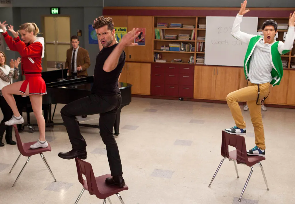 Heather Morris, Ricky Martin, and Harry Shum Jr. dancing on chairs in a scene from Glee.