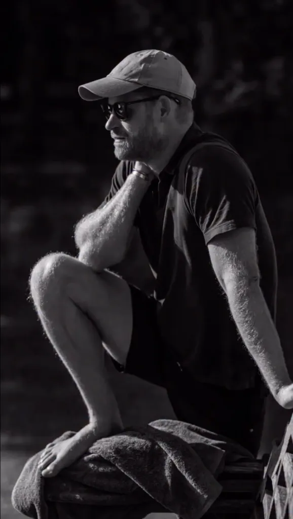 Prince Harry in a baseball cap, sunglasses, t-shirt, and shorts, crouching with one foot on a towel.
