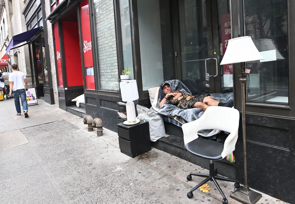 A homeless man lies on a bed made of blankets and pillows on the street next to a building, with a desk lamp and office chair nearby.