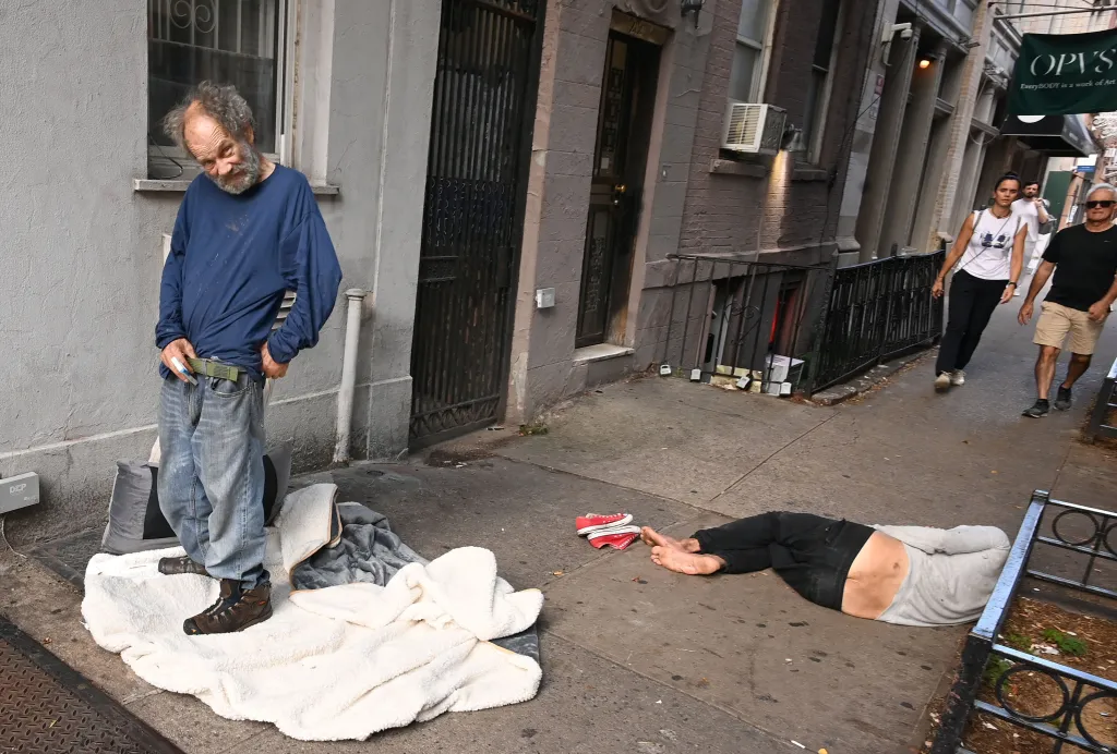 Homeless man, Louis, 71, standing on a sidewalk on Sullivan Street, with another person lying on the ground next to him and three other people walking by in the background.